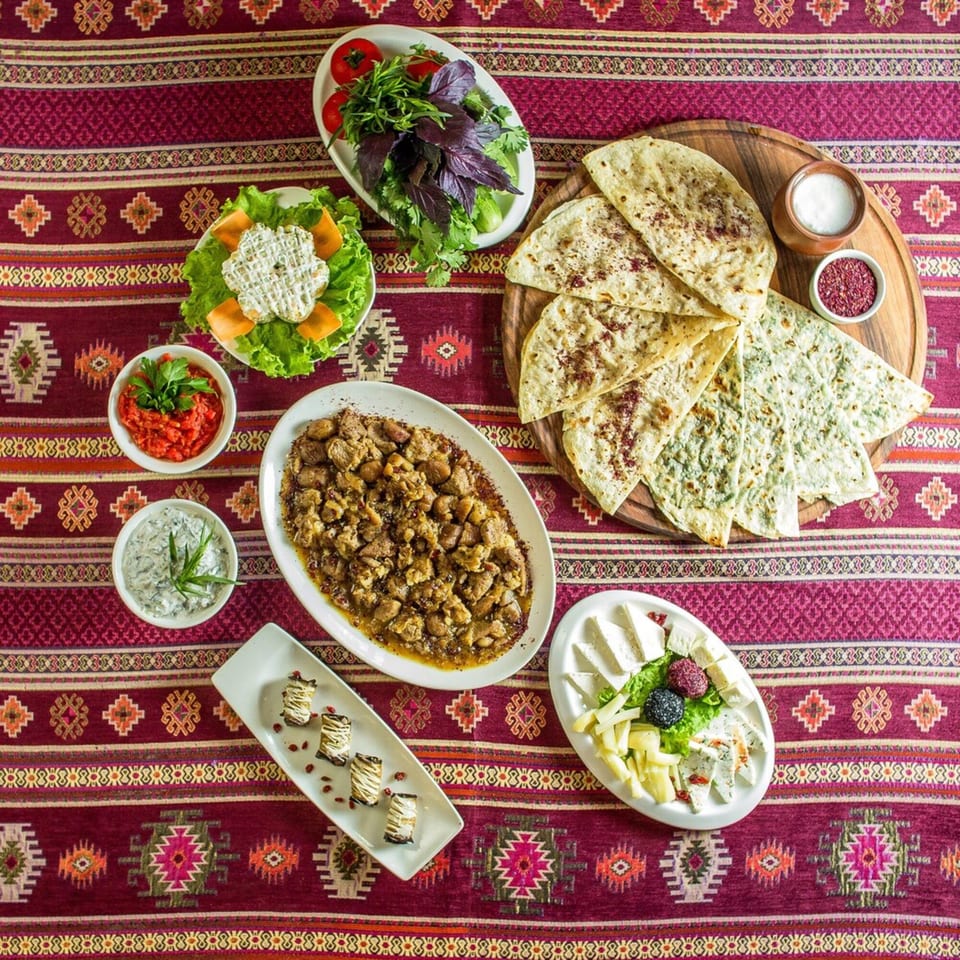 A vibrant Lebanese feast on a patterned red tablecloth, featuring meat, flatbreads, cheeses, and salads.